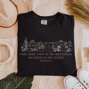 Puede incluir: Camiseta negra con un dise&ntilde;o de paisaje des&eacute;rtico blanco y el texto "I WILL MAKE A WAY IN THE WILDERNESS AND RIVERS IN THE DESERT ISAIAH 43:19". La camiseta es de cuello redondo y tiene una etiqueta que dice "COMFORT COLORS".