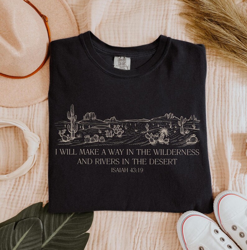 Puede incluir: Camiseta negra con un dise&ntilde;o de paisaje des&eacute;rtico blanco y el texto "I WILL MAKE A WAY IN THE WILDERNESS AND RIVERS IN THE DESERT ISAIAH 43:19". La camiseta es de cuello redondo y tiene una etiqueta que dice "COMFORT COLORS".