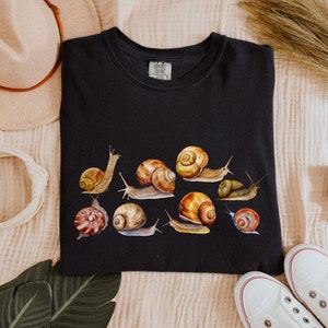 May include: Black t-shirt with a graphic print of multiple snails in various colors and patterns. The text "Comfort Colors" is visible on the tag.