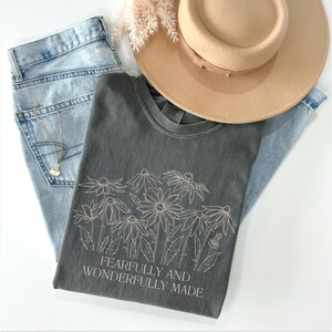 May include: A charcoal grey t-shirt with a white line drawing of flowers and the text "Fearfully and Wonderfully Made".