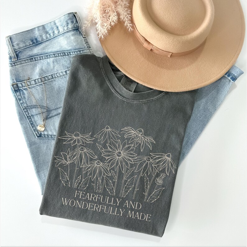 May include: A charcoal grey t-shirt with a white line drawing of flowers and the text "Fearfully and Wonderfully Made".