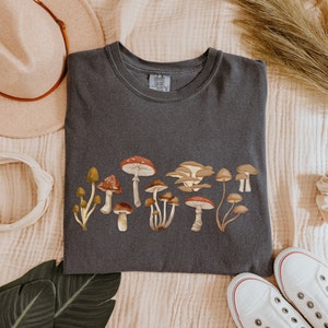 May include: A dark gray t-shirt with a graphic of various mushrooms in shades of brown, red, and yellow. The text "Comfy Colors" is visible on the tag.
