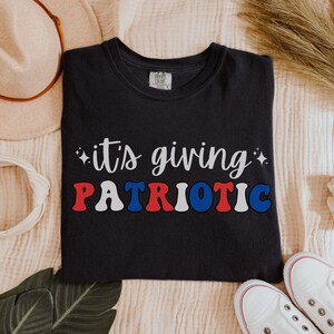 Pu&ograve; includere: T-shirt blu navy con la scritta "it's giving PATRIOTIC" in bianco, rosso e blu. La maglietta ha un girocollo e maniche corte. Il design &egrave; centrato sul davanti.