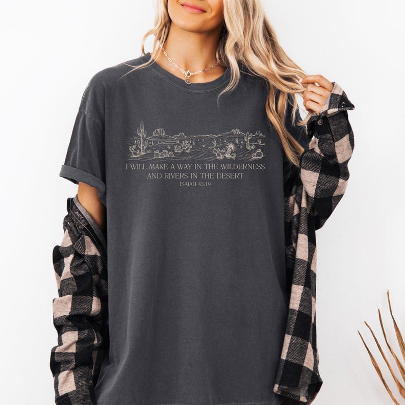 Puede incluir: Camiseta gris oscuro con un dise&ntilde;o gr&aacute;fico de paisaje des&eacute;rtico, incluyendo cactus, y el texto "I WILL MAKE A WAY IN THE WILDERNESS AND RIVERS IN THE DESERT ISAIAH 43:19". La camiseta tiene mangas cortas y un corte relajado.