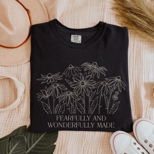 May include: A black t-shirt with a white outline of flowers and the text "Fearfully and Wonderfully Made".