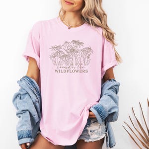 May include: A pink t-shirt with a brown graphic of wildflowers and the text "consider the wildflowers".