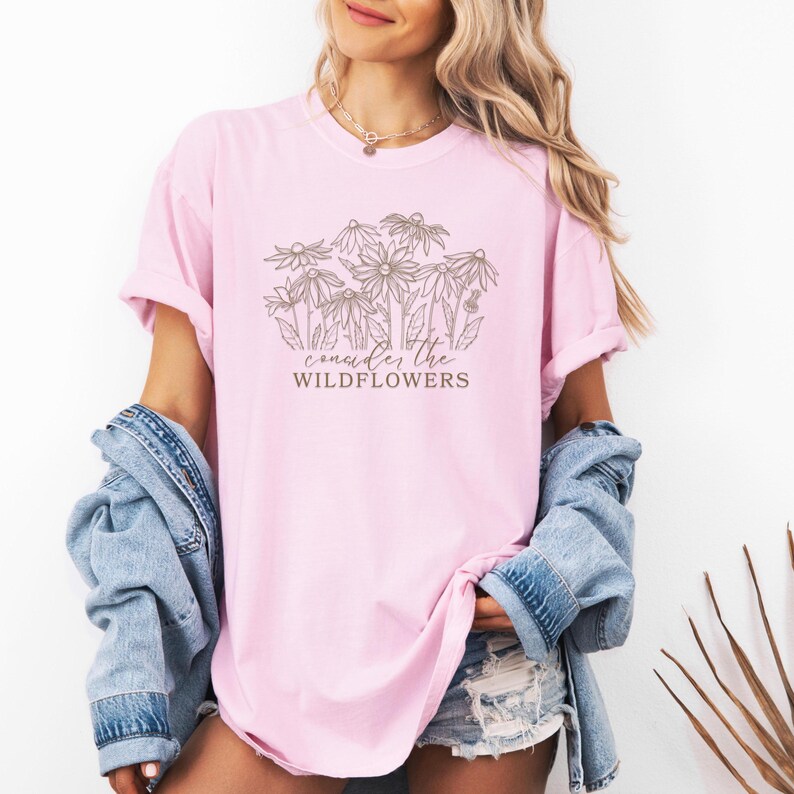 May include: A pink t-shirt with a brown graphic of wildflowers and the text "consider the wildflowers".