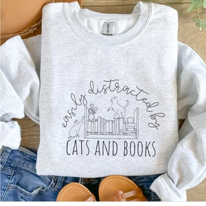 May include: A gray sweatshirt with a black graphic design of two cats on a bookshelf with the text "easily distracted by cats and books".
