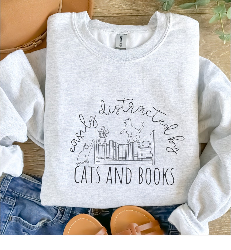 May include: A gray sweatshirt with a black graphic design of two cats on a bookshelf with the text "easily distracted by cats and books".