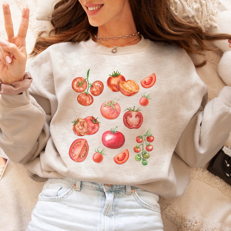 May include: A light gray sweatshirt with a watercolor design of red tomatoes. The tomatoes are in various shapes and sizes, some are whole and some are sliced.