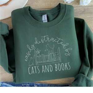 May include: A dark green sweatshirt with a white graphic design that says "easily distracted by cats and books". The graphic features a bookshelf with cats and books.