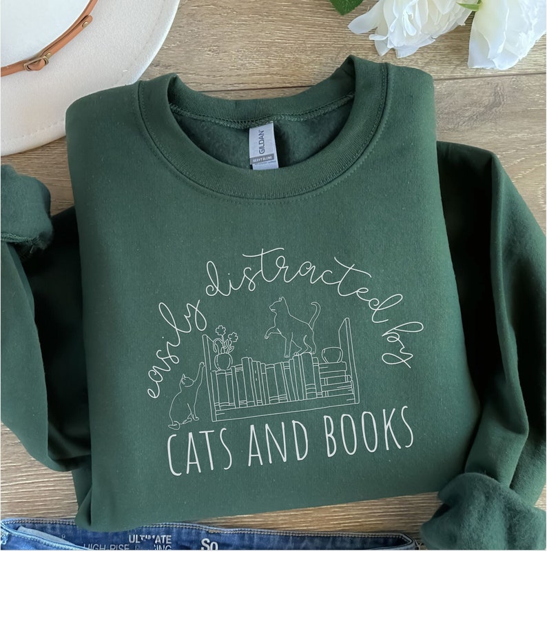 May include: A dark green sweatshirt with a white graphic design that says "easily distracted by cats and books". The graphic features a bookshelf with cats and books.