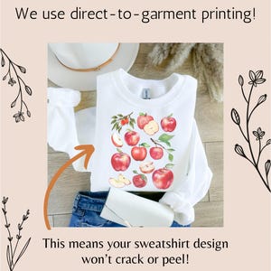 Pu&ograve; includere: Felpa bianca con un design di mele rosse. Il testo "We use direct-to-garment printing!" &egrave; nella parte superiore dell'immagine. Il testo "This means your sweatshirt design won't crack or peel!" &egrave; nella parte inferiore dell'immagine.