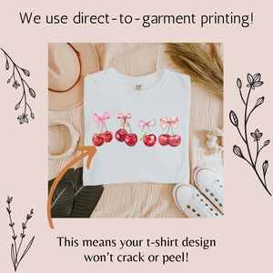 Puede incluir: Camiseta blanca con un dise&ntilde;o de cinco cerezas rojas con lazos rosas. El texto "We use direct-to-garment printing!" y "This means your t-shirt design won't crack or peel!" es visible en la imagen.
