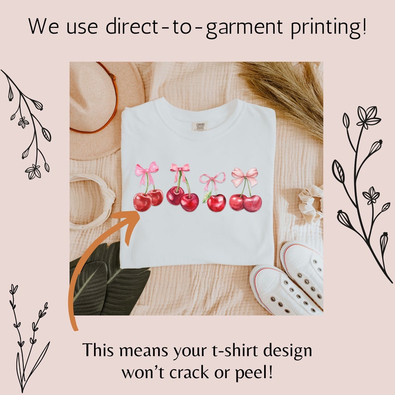 Puede incluir: Camiseta blanca con un dise&ntilde;o de cinco cerezas rojas con lazos rosas. El texto "We use direct-to-garment printing!" y "This means your t-shirt design won't crack or peel!" es visible en la imagen.