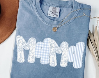 May include: A blue Comfort Colors t-shirt with the word "Mama" appliqued across the front. The letters are a mix of floral, gingham, and striped patterns. A gold necklace with a pendant rests on the shirt.