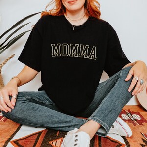 May include: A black t-shirt with the word "MOMMA" printed in gold on the front. The shirt is being worn by a person sitting on a rug.
