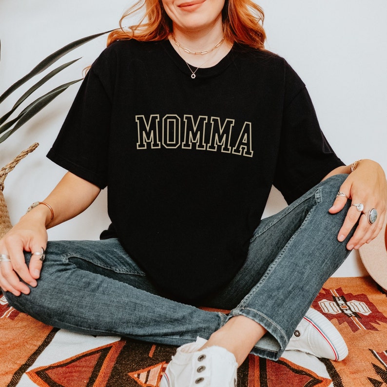 May include: A black t-shirt with the word "MOMMA" printed in gold on the front. The shirt is being worn by a person sitting on a rug.