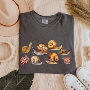 May include: A dark gray t-shirt with a print of various snails in different colors and sizes. The snails are arranged in a row across the shirt. The shirt has a crew neck and short sleeves.