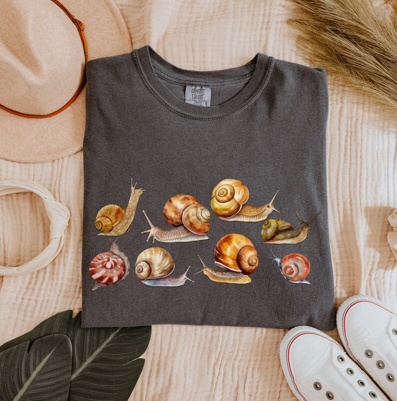 May include: A dark gray t-shirt with a print of various snails in different colors and sizes. The snails are arranged in a row across the shirt. The shirt has a crew neck and short sleeves.