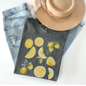 Pu&ograve; includere: Una maglietta grigia antracite con una stampa di limone. I limoni sono gialli con foglie verdi e sono disposti in modo casuale. La maglietta &egrave; appoggiata su un paio di jeans blu e un cappello marrone.