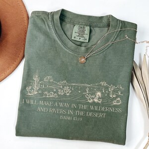 Puede incluir: Camiseta verde oliva Comfort Colors con un gr&aacute;fico de paisaje des&eacute;rtico y el texto "I WILL MAKE A WAY IN THE WILDERNESS AND RIVERS IN THE DESERT ISAIAH 43:19". Un collar dorado con un colgante est&aacute; sobre la camiseta. Un sombrero marr&oacute;n est&aacute; en el fondo.