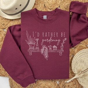 Peut inclure: Un sweat-shirt bordeaux avec le texte "I'd rather be gardening" en blanc. Le texte est accompagn&eacute; d'un dessin au trait d'une &eacute;tag&egrave;re avec des plantes en pot.