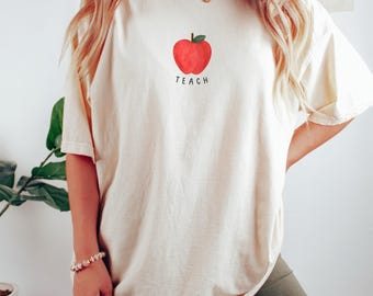May include: A white oversized t-shirt with a red apple graphic and the word "TEACH" printed below it.