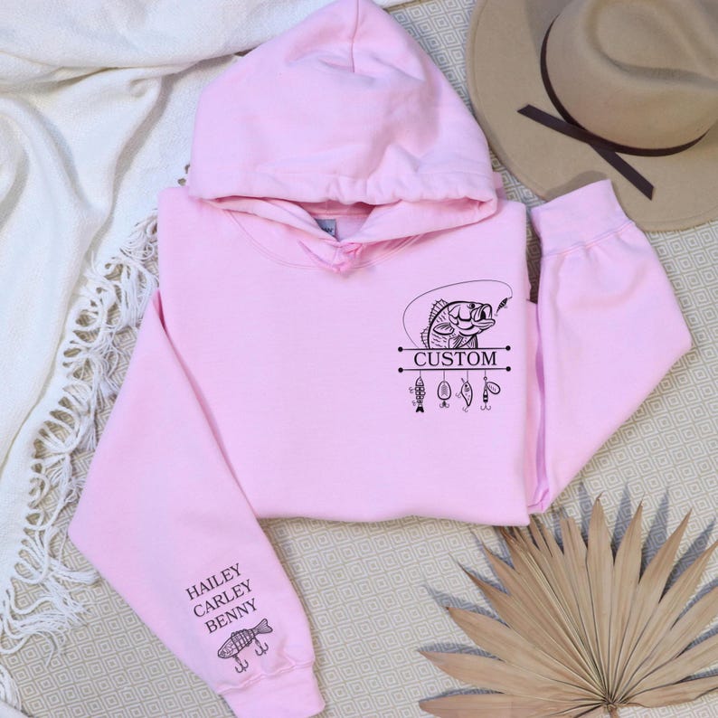 Pode incluir: Camisola de capuz rosa com um gr&aacute;fico preto de um peixe numa linha de pesca com a palavra "CUSTOM" e iscas de pesca. A manga tem os nomes "HAILEY, CARLEY, BENNY" e um gr&aacute;fico de peixe.