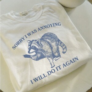 May include: White t-shirt with a blue raccoon graphic and the text "Sorry I was annoying I will do it again".