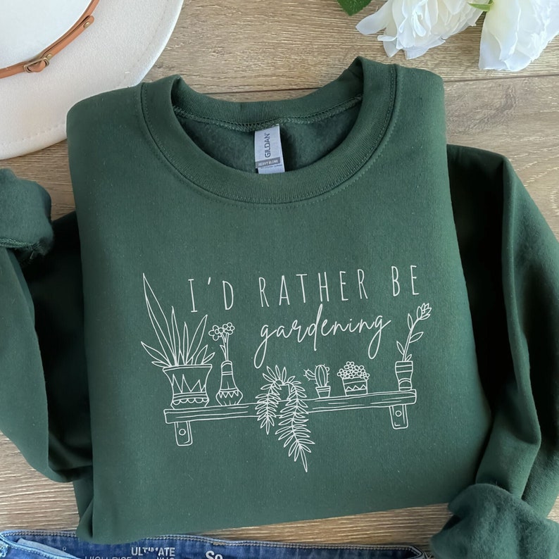 Peut inclure: Un sweat-shirt vert fonc&eacute; avec le texte "I'd rather be gardening" en lettres blanches. Le texte est accompagn&eacute; d'un dessin au trait d'une &eacute;tag&egrave;re avec des plantes en pot.