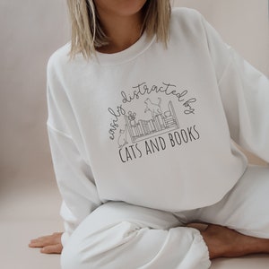 May include: White sweatshirt with a black graphic design that says "easily distracted by cats and books". The graphic features a bookshelf with cats and plants.
