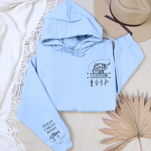 Pode incluir: Camisola de capuz azul claro com um gr&aacute;fico preto e branco de um peixe numa vara de pesca com a palavra "CUSTOM" e iscas de pesca. A manga esquerda tem os nomes "HAILEY CARLEY BENNY" e um pequeno gr&aacute;fico de peixe.