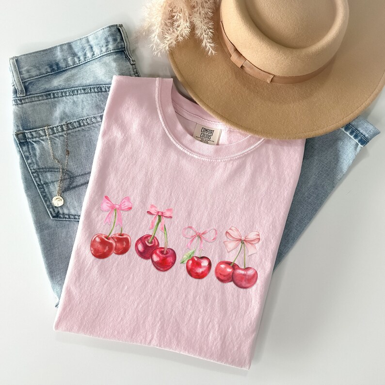 Puede incluir: Camiseta rosa con un gr&aacute;fico de cinco cerezas rojas con lazos rosas en los tallos. La camiseta tiene una etiqueta "Comfy Colors" en la parte inferior.