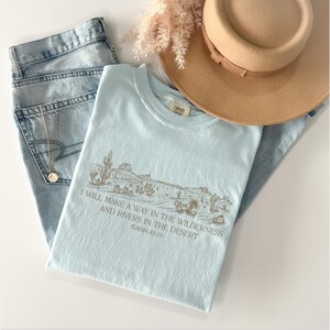 Puede incluir: Una camiseta azul claro con un gr&aacute;fico de paisaje des&eacute;rtico y el texto "I WILL MAKE A WAY IN THE WILDERNESS AND RIVERS IN THE DESERT ISAIAH 43:19". La camiseta se combina con vaqueros de lavado claro, un sombrero beige y un collar dorado.