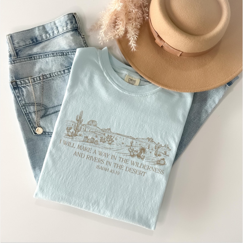 Puede incluir: Una camiseta azul claro con un gr&aacute;fico de paisaje des&eacute;rtico y el texto "I WILL MAKE A WAY IN THE WILDERNESS AND RIVERS IN THE DESERT ISAIAH 43:19". La camiseta se combina con vaqueros de lavado claro, un sombrero beige y un collar dorado.