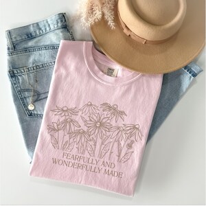May include: A pink t-shirt with a brown outline of flowers and the text "Fearfully and Wonderfully Made".