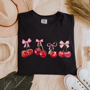 Puede incluir: Camiseta negra con un gr&aacute;fico de cinco cerezas rojas con lazos rosas. Las cerezas est&aacute;n dispuestas en una fila a trav&eacute;s de la parte delantera de la camiseta.