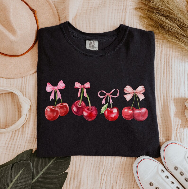 Puede incluir: Camiseta negra con un gr&aacute;fico de cinco cerezas rojas con lazos rosas. Las cerezas est&aacute;n dispuestas en una fila a trav&eacute;s de la parte delantera de la camiseta.
