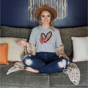 May include: A woman wearing a grey t-shirt with a pink and black leopard print heart design and a red heart. The shirt has the text "Love" on it. She is sitting on a couch with a brown hat on her head.