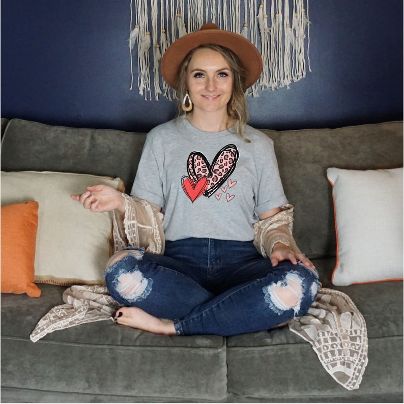 May include: A woman wearing a grey t-shirt with a pink and black leopard print heart design and a red heart. The shirt has the text "Love" on it. She is sitting on a couch with a brown hat on her head.