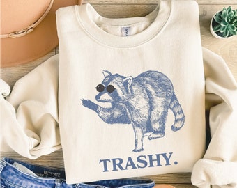 May include: A cream-colored sweatshirt with a blue illustration of a raccoon wearing sunglasses and the word "TRASHY." printed below.