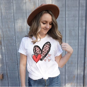 May include: White t-shirt with a graphic of two hearts. The larger heart is pink with a leopard print and the smaller heart is red. There are also smaller pink hearts scattered around the larger hearts.