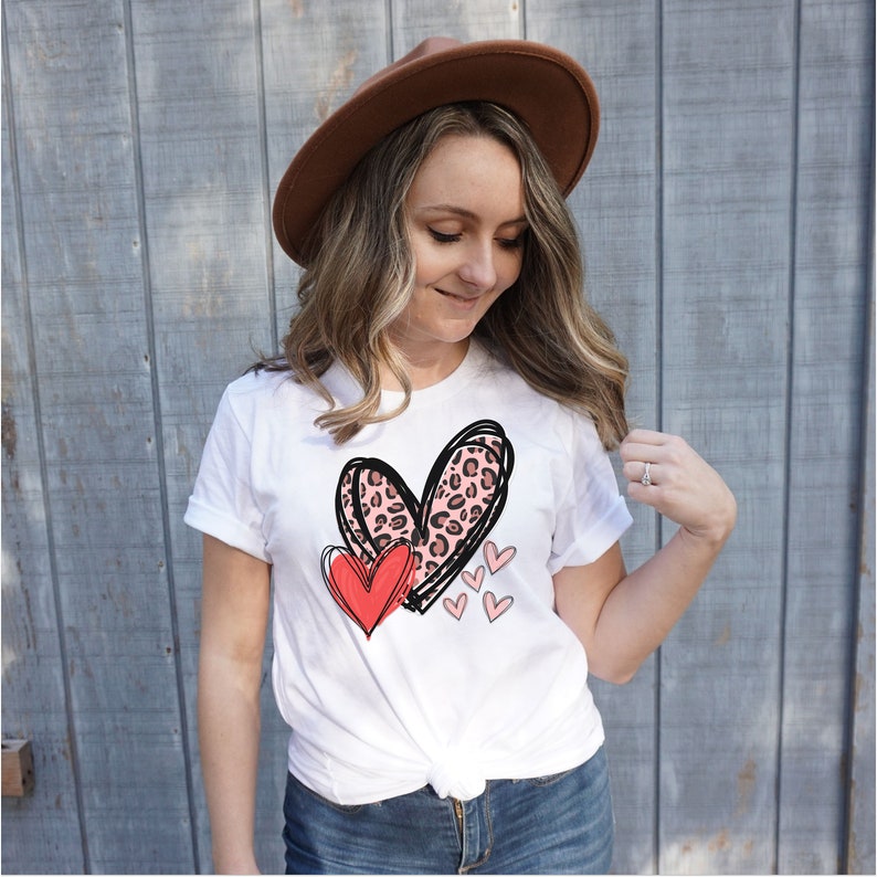 May include: White t-shirt with a graphic of two hearts. The larger heart is pink with a leopard print and the smaller heart is red. There are also smaller pink hearts scattered around the larger hearts.