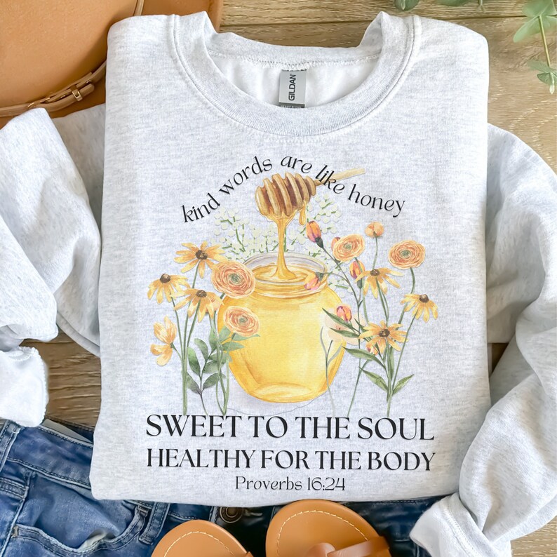 Puede incluir: Una sudadera gris con un tarro de miel y flores impresas en la parte delantera. El texto dice "Kind words are like honey, Sweet to the soul, Healthy for the body, Proverbs 16:24".