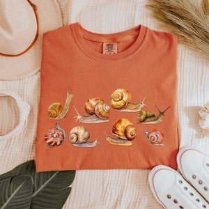 May include: A rust-colored t-shirt with a graphic of multiple snails in various poses. The snails are painted in shades of brown, yellow, and white.