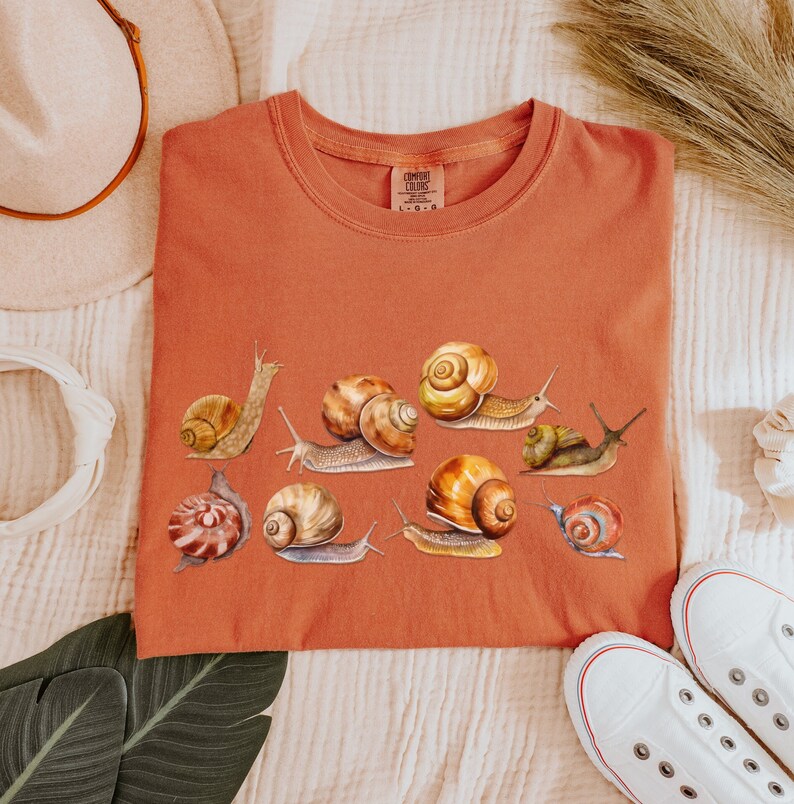 May include: A rust-colored t-shirt with a graphic of multiple snails in various poses. The snails are painted in shades of brown, yellow, and white.