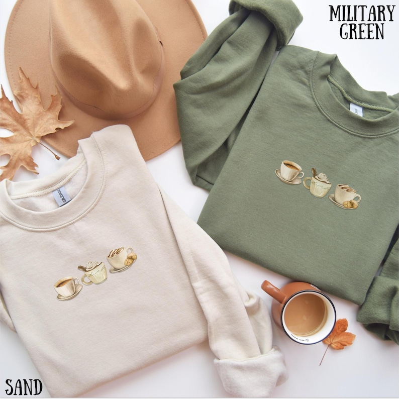 May include: Two sweatshirts, one sand-colored and one military green, each embroidered with three coffee cups. A brown hat and a cup of coffee are also visible. The word "SAND" is printed at the bottom left, and "MILITARY GREEN" is at the top right.