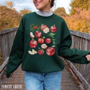 Pu&ograve; includere: Una felpa verde bosco con una stampa grafica di mele rosse e fette di mela. Le mele sono disposte a grappolo e alcune sono tagliate a met&agrave; per mostrare l'interno.