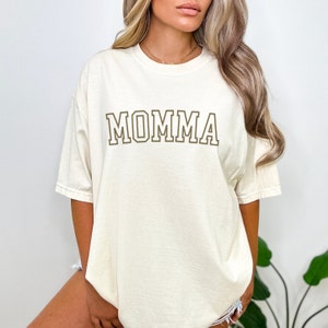 May include: A white oversized t-shirt with the word "MOMMA" printed in brown on the front.